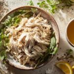 A ceramic bowl filled with healthy shredded chicken recipes garnished with fresh herbs and lemon wedges.