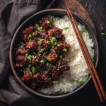 A ceramic bowl filled with sticky, glazed bourbon chicken recipe served over white rice and garnished with green scallions.